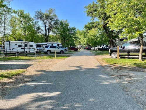 A gravel road lined with RVs and trees in a sunny campground setting.