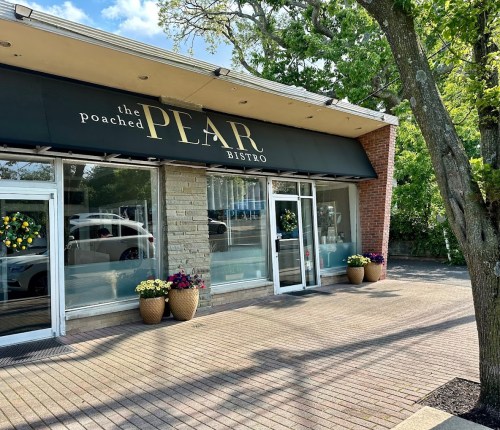 Exterior of The Poached Pear Bistro, featuring large windows, a black awning, and flower pots on a sunny day.