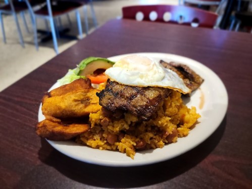 A plate of rice and beans topped with grilled meat, a fried egg, plantains, and a side of lettuce and tomato.