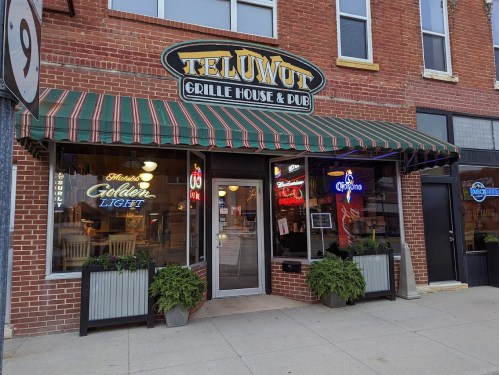Exterior of Teluwut Grille House & Pub, featuring a striped awning, neon signs, and potted plants by the entrance.