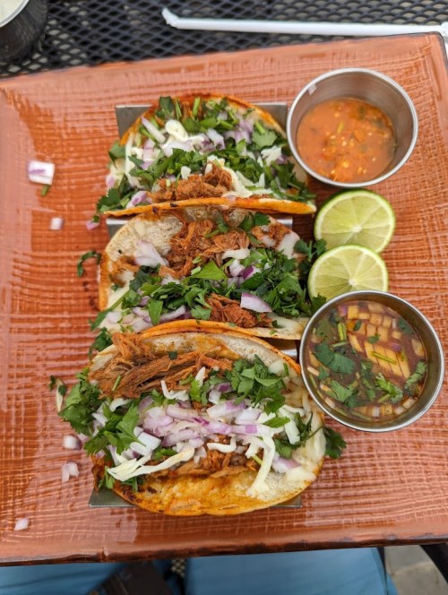 Three tacos topped with shredded meat, onions, and cilantro, served with lime wedges and two small bowls of sauce.