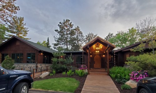 A rustic wooden building with a welcoming entrance, surrounded by greenery and colorful flowers.