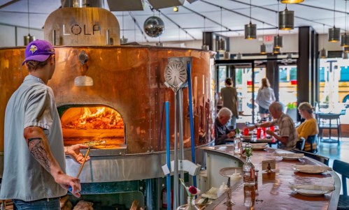 A chef prepares food in a large, copper wood-fired oven at a bustling restaurant with diners enjoying their meals.