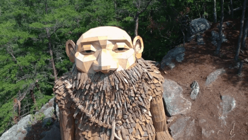 A large wooden sculpture of a bearded figure stands among trees and rocks, showcasing intricate craftsmanship.