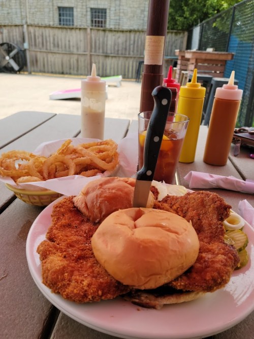 A plate with a large fried chicken sandwich, two buns, pickles, onion rings, and condiments in the background.