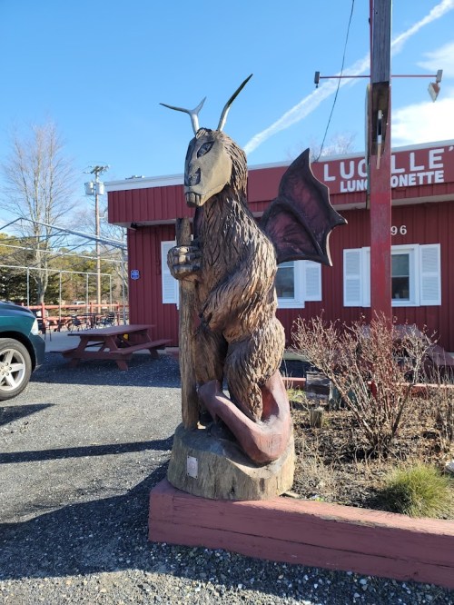 A wooden sculpture of a creature with goat features, wings, and a staff, standing outside a red building.