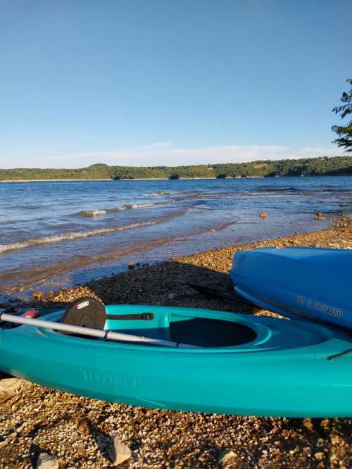 A turquoise kayak rests on a pebbly shore by a calm lake, with trees and hills in the background under a clear blue sky.