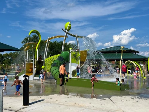 A vibrant splash pad with water features, children playing, and families enjoying a sunny day at the park.