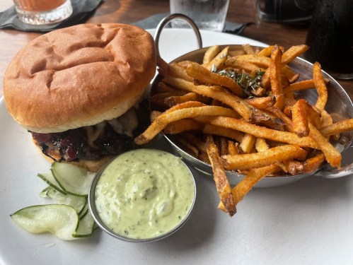 A burger with a bun, topped with sauce, served with a side of crispy fries and a small bowl of green dipping sauce.
