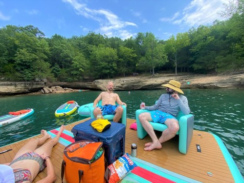 Two men relax on a dock by the water, surrounded by trees, with snacks and drinks nearby. A woman is partially visible.
