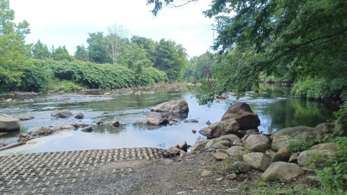 A serene river scene with rocks, greenery, and a calm water surface under a clear sky.