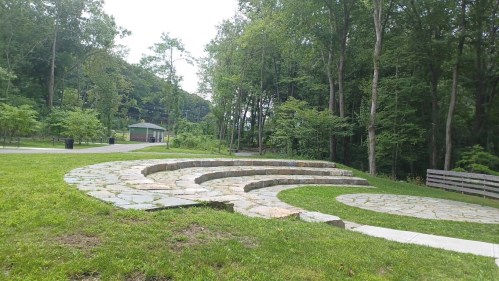A stone amphitheater in a wooded park, surrounded by greenery and a path leading to a nearby gazebo.