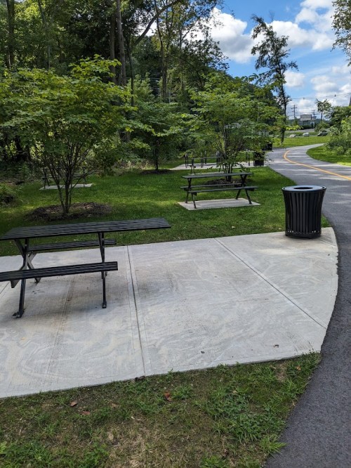 A park path with picnic tables and a trash can, surrounded by greenery and trees under a blue sky.