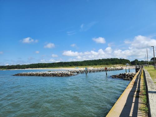 A serene waterfront scene with a rocky jetty, calm water, and a sandy beach under a blue sky with scattered clouds.