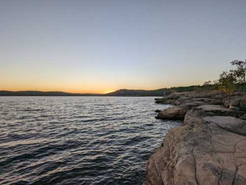 A serene lakeside view at sunset, with calm waters and rocky shores under a clear sky.