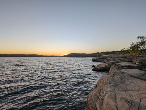 A serene lakeside view at sunset, with calm waters and rocky shores under a clear sky.