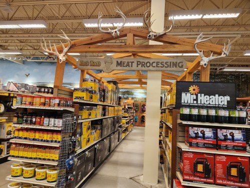 A store aisle labeled "Meat Processing" with shelves of supplies and deer antlers overhead.