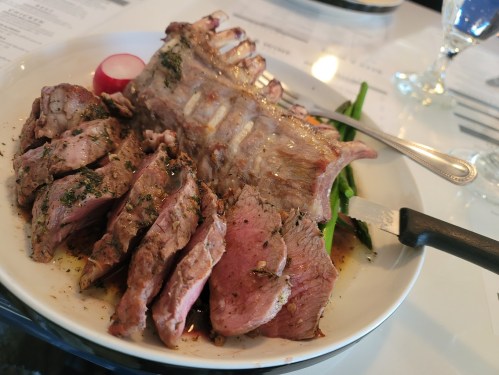 A plate of sliced roasted meat with a rack of ribs, garnished with herbs and served with asparagus and a radish.