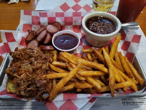 A platter featuring pulled pork on bread, fries, sausage slices, and a bowl of baked beans, served on a checkered paper.