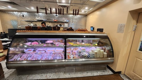 A display case filled with various meats and prepared foods in a restaurant, with a staff member in the background.