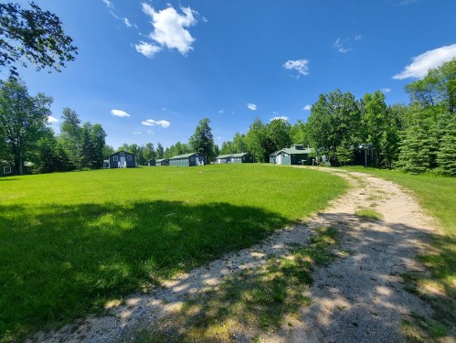 A grassy area with a dirt path leading to green cabins under a blue sky with scattered clouds.