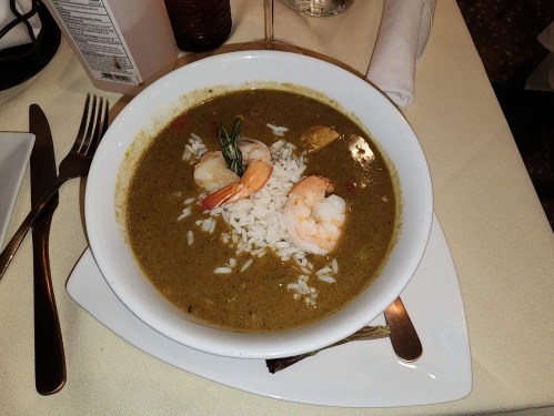 A bowl of green gumbo with shrimp and rice, served on a white plate with utensils beside it.