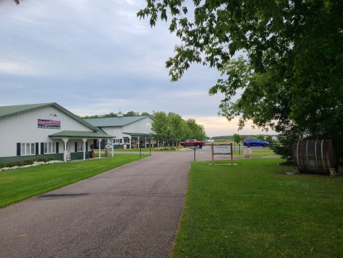 A scenic view of a driveway leading to a building with green landscaping and parked cars under a cloudy sky.