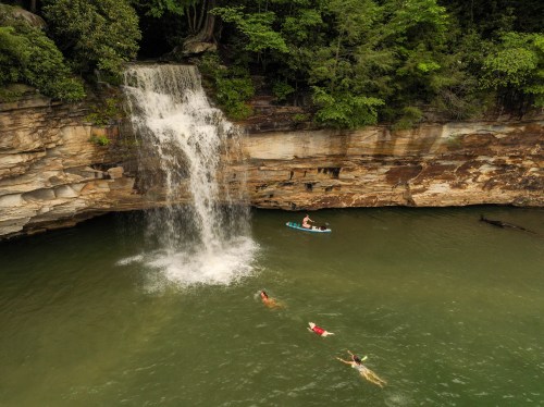 A waterfall cascades into a green pool, with people swimming and a kayak nearby, surrounded by lush trees.