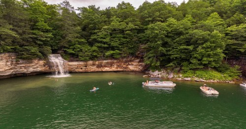 A serene lake surrounded by lush greenery, featuring a waterfall and people enjoying boats and swimming.