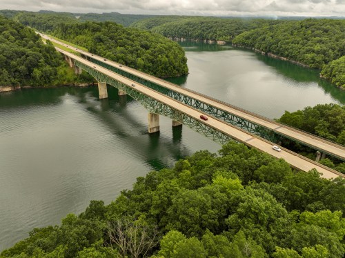 Aerial view of a bridge spanning a river, surrounded by lush green trees and rolling hills under a cloudy sky.