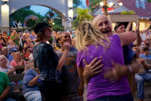 A joyful crowd claps and celebrates as two people embrace in a lively outdoor setting.