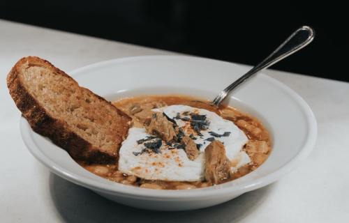 A bowl of soup with meat, topped with a poached egg and herbs, served with a slice of brown bread on the side.