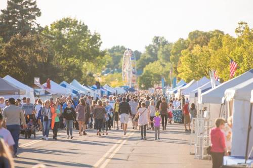 A bustling street fair with crowds, vendor tents, and a ferris wheel in the background under a sunny sky.