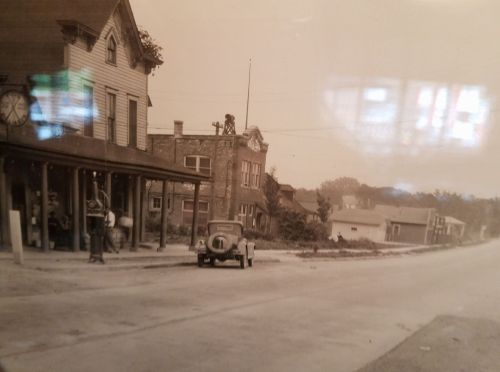 A vintage street scene featuring a clock, buildings, and an old car parked along a quiet road.