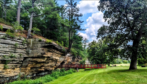 A scenic view of a rocky cliff with trees, a grassy area, and a red wooden bridge under a partly cloudy sky.