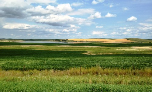 A scenic landscape featuring green fields, a calm lake, and rolling hills under a partly cloudy sky.