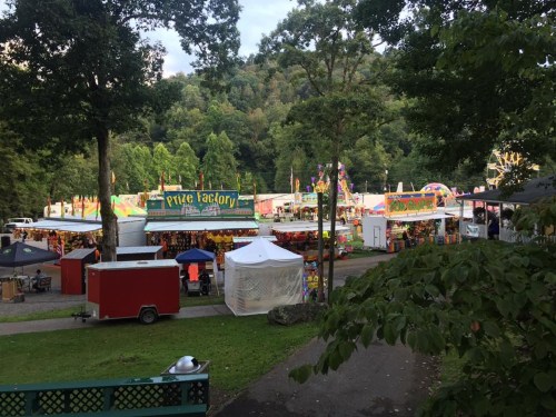 A vibrant fairground with colorful booths and attractions, surrounded by trees and hills in the background.
