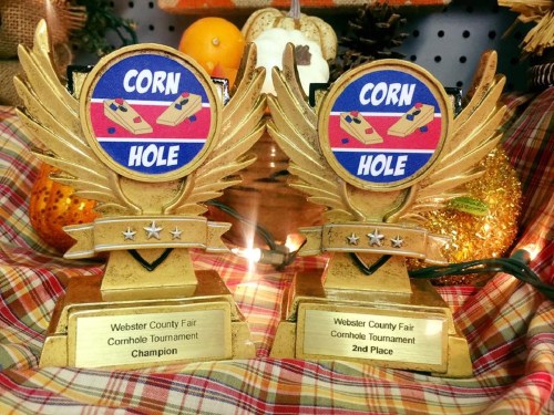 Two trophies with gold wings, labeled "Champion" and "2nd Place," for a Cornhole Tournament at the Webster County Fair.