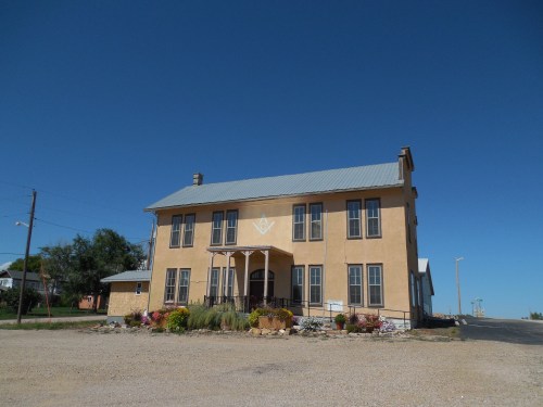 A two-story yellow building with a metal roof, featuring a front porch and surrounded by flowers against a clear blue sky.