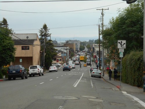 A street view showing cars, buildings, and pedestrians in a small town, with a cloudy sky in the background.