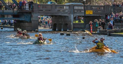 A group of kayakers paddles on a river, with a crowd watching from a bridge and riverbank in the background.