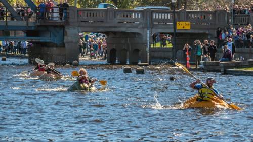 Four kayakers paddle through a river, with a crowd watching from a nearby bridge and bank on a sunny day.