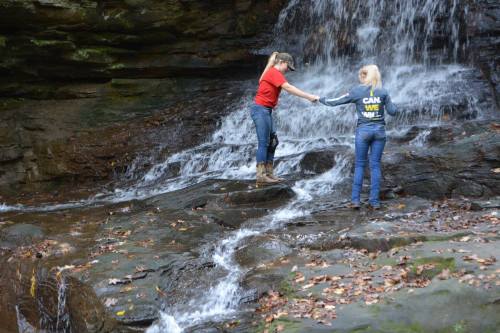 Two people stand on rocks near a waterfall, reaching out to each other amidst flowing water and autumn leaves.