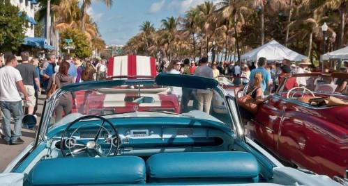 A view from inside a classic convertible, overlooking a crowded street filled with people and vintage cars under palm trees.