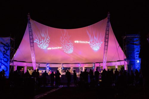 A large tent illuminated with colorful lights, with a crowd gathered in front during a nighttime event.