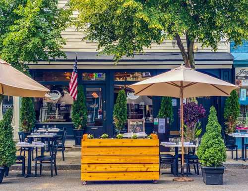 A cozy outdoor dining area with umbrellas, a wooden planter, and a restaurant facade featuring greenery and an American flag.