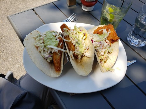 Three fish tacos on a plate, topped with slaw and avocado sauce, served outdoors with drinks in the background.