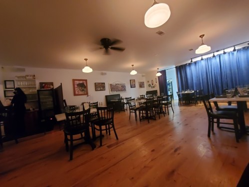 A spacious restaurant interior with wooden floors, black chairs, and blue curtains, featuring a few tables and a staff member.