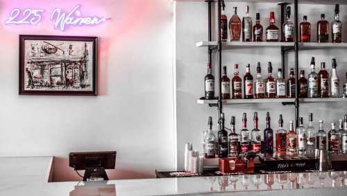 A stylish bar with a neon sign, displaying various bottles of liquor on shelves against a white wall.