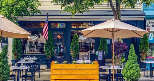 Outdoor dining area of a restaurant with tables, umbrellas, and greenery, featuring an American flag and a welcoming entrance.